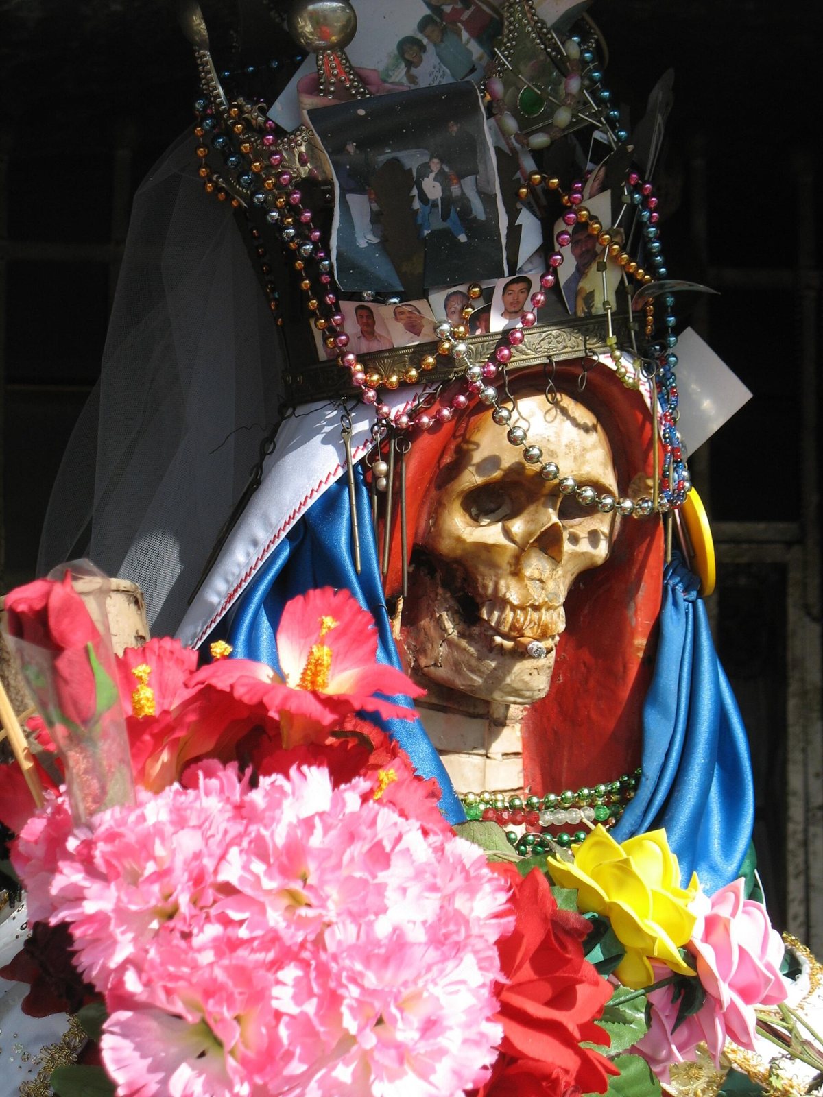 A Santa Muerte figure in a red and blue robe, with strings of beads and small photographs pinned to her crown, surrounded by pink, red and yellow artificial flowers