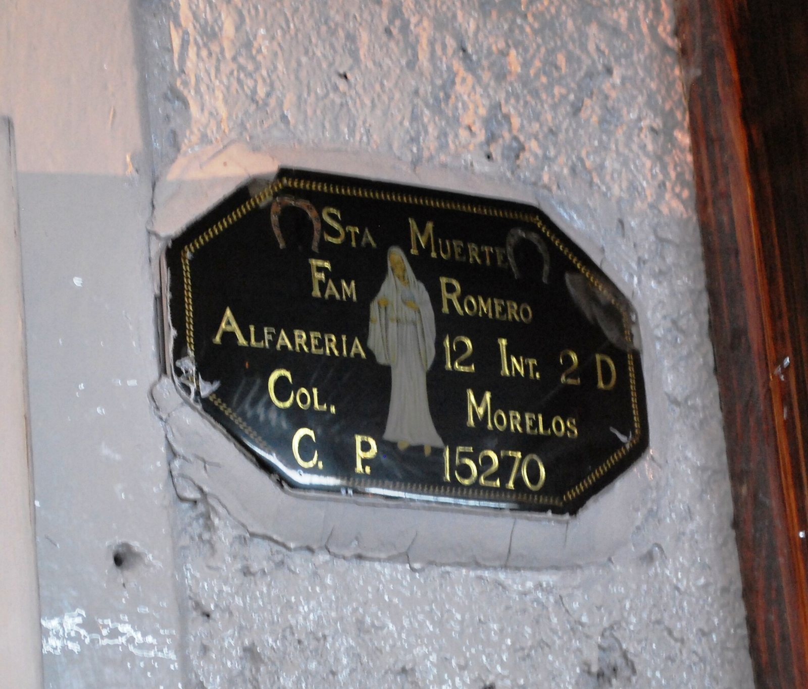 A black plaque on a gray wall reading Sta Muerte, Fam Romero, Alfareria 12, Col. Morelos, with a small robed figure painted in the center