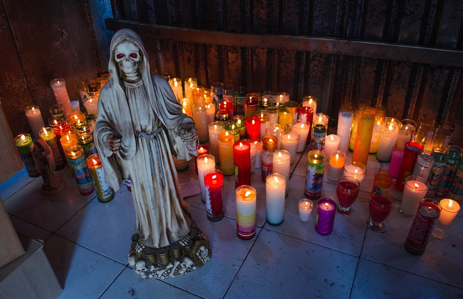 A pale robed Santa Muerte statue standing on a tiled floor beside dozens of lit candles in white, red, yellow, purple and green glass jars