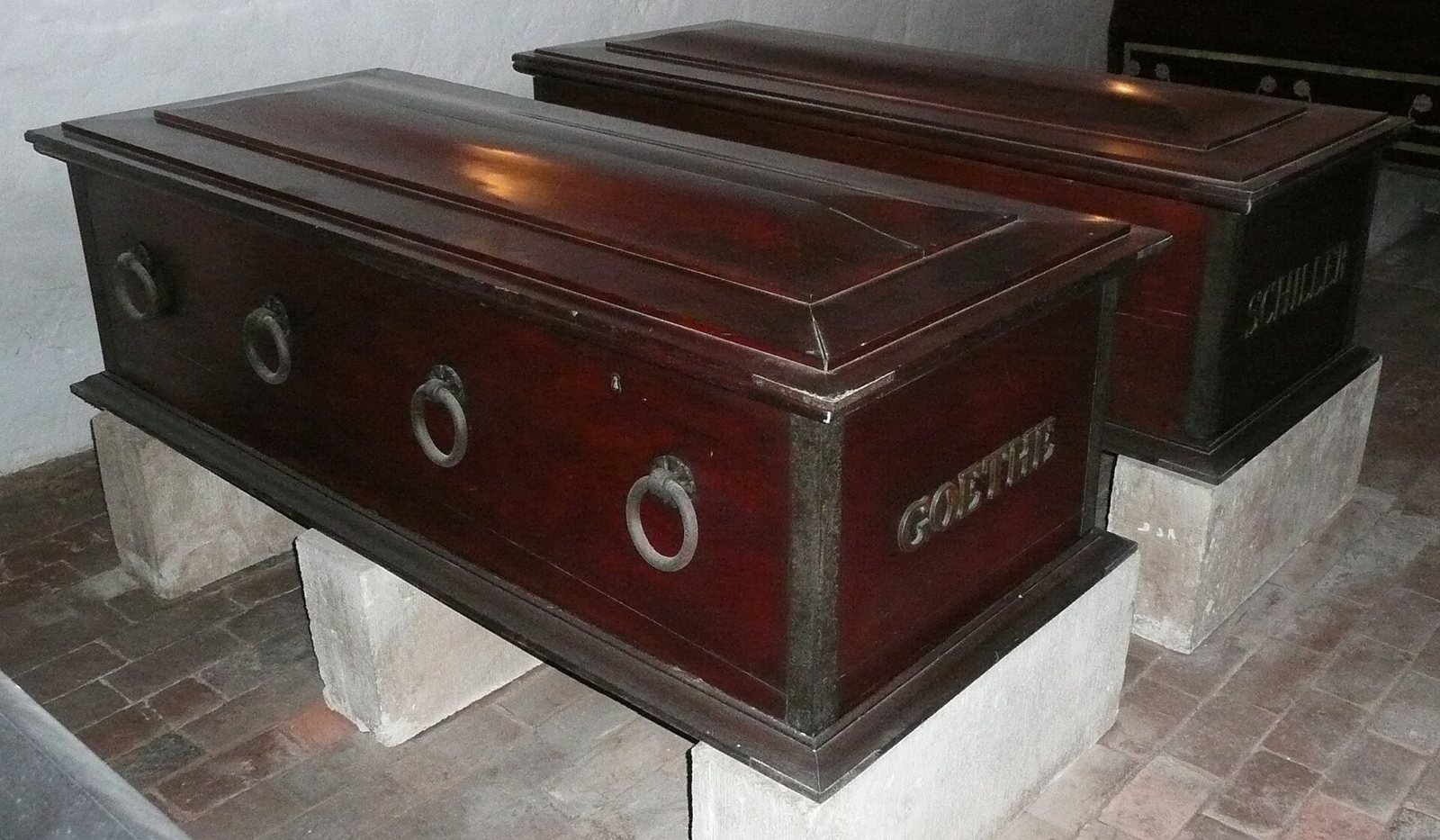 Two dark wooden sarcophagi with raised lids on pale stone plinths in the Fürstengruft in Weimar, the nearer one lettered GOETHE and the one behind it SCHILLER