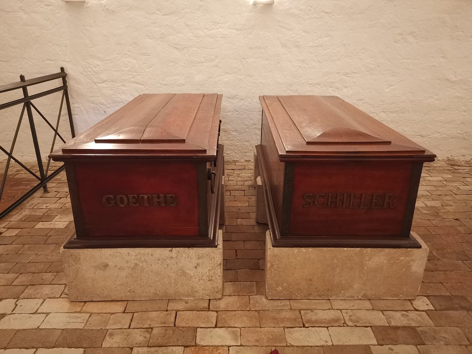 Two reddish-brown wooden sarcophagi on pale stone bases against a white wall in the Fürstengruft, lettered GOETHE on the left and SCHILLER on the right, on a brick floor