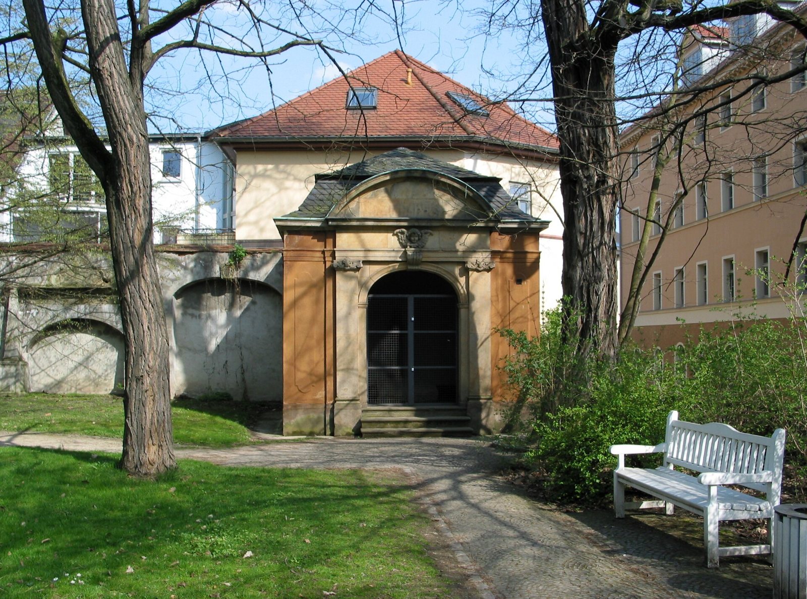 The Kassengewölbe on the Jakobskirchhof in Weimar, a small yellow-ochre stone building with an arched doorway and a red-tiled roof, seen between bare trees, with a bench in the foreground