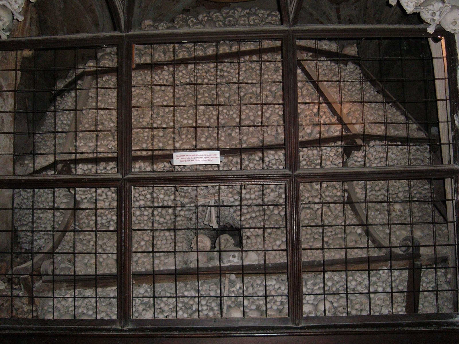 A tall pile of skulls and bones stacked in a vaulted niche behind a dark metal grille, with a small information plaque on the grille