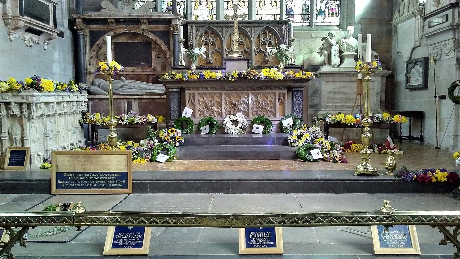 The chancel of Holy Trinity Church, Stratford-upon-Avon, with flowers laid along the altar steps and signs marking the graves of Shakespeare and his family in the floor