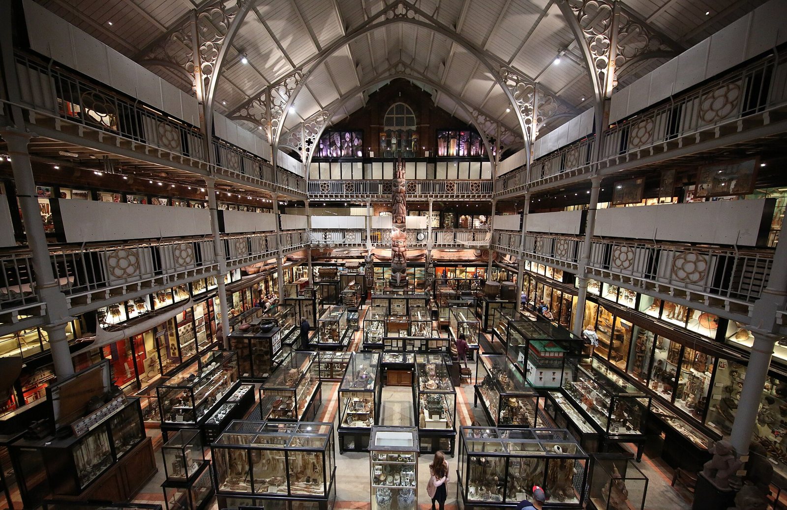 The interior of the Pitt Rivers Museum seen from an upper gallery: rows of glass display cases on the ground floor beneath tiered iron balconies and a high glass and iron roof