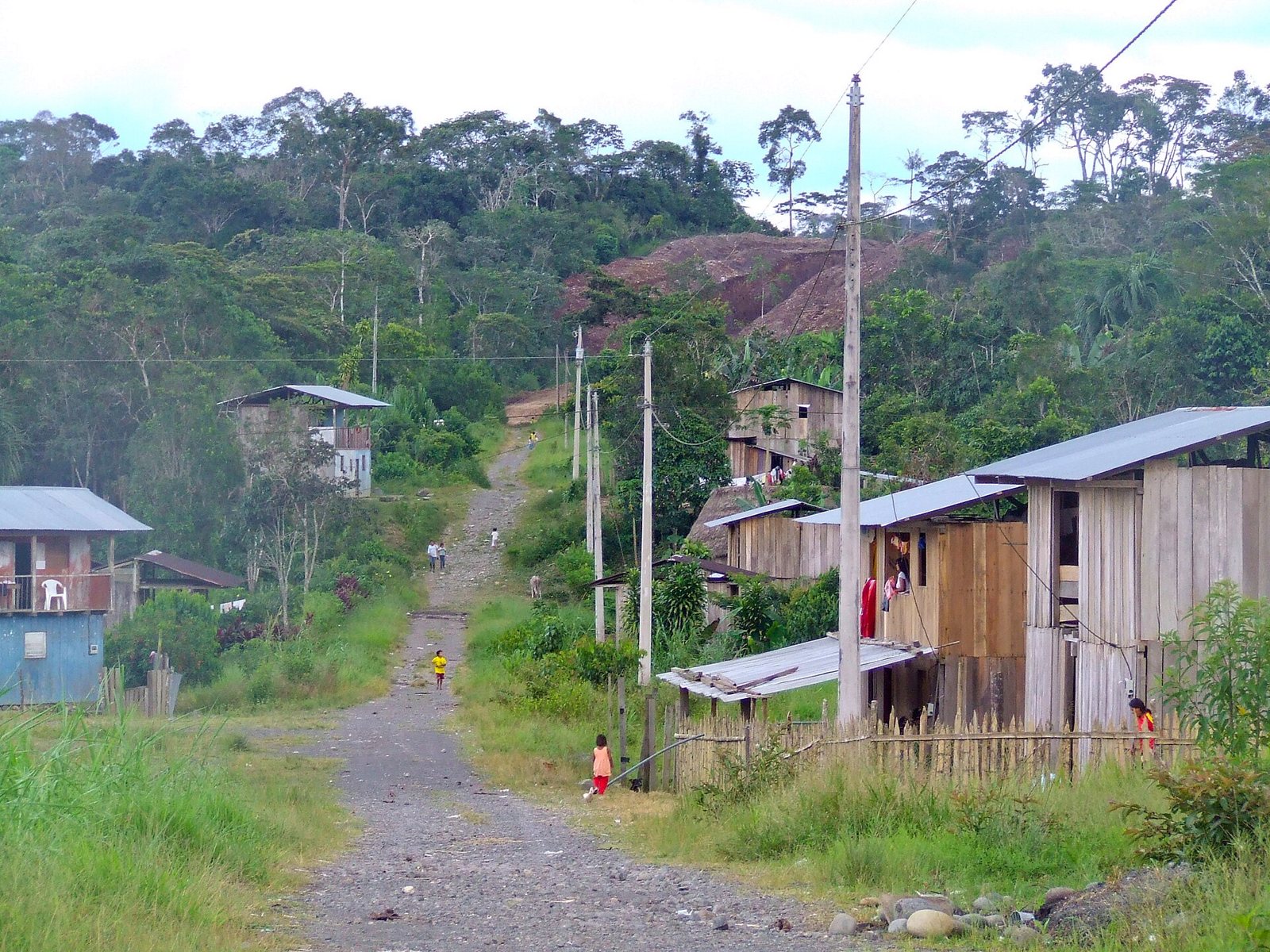 A gravel road running up through a Shuar village on a forested hillside, lined with wooden houses on stilts with metal roofs and tall power poles