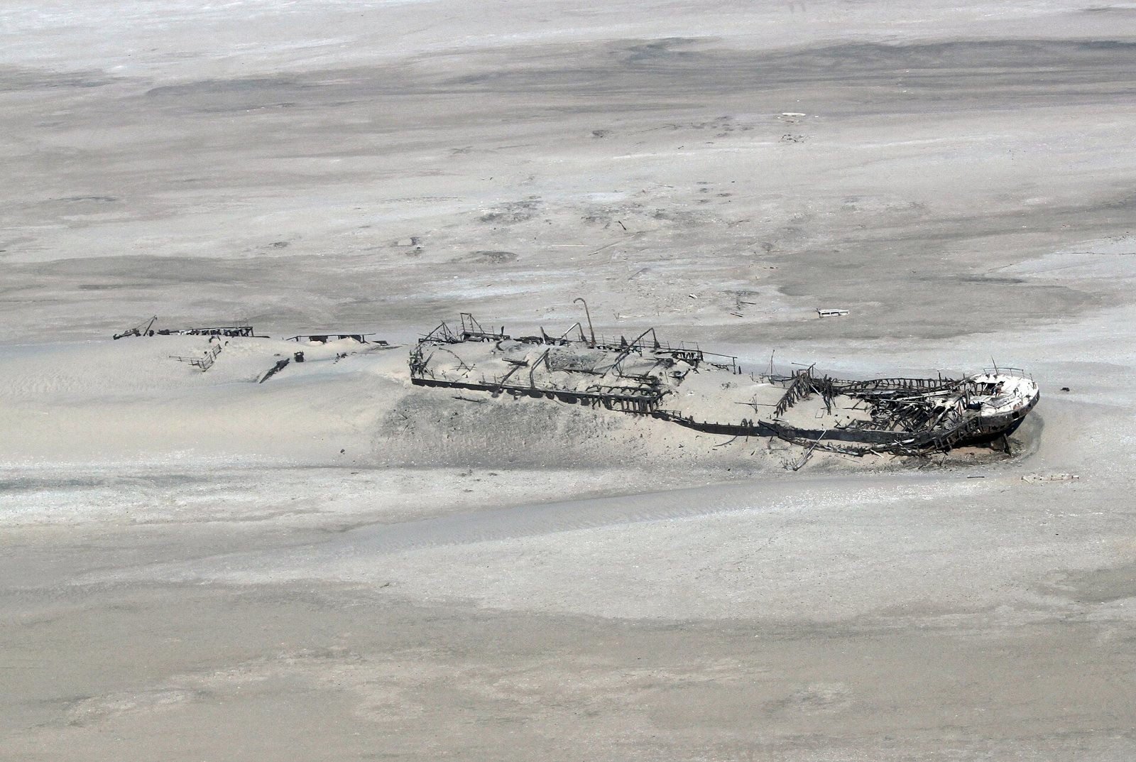 The skeletal remains of the Eduard Bohlen seen from above, a dark broken hull and ribs half buried in pale sand with sea and shoreline behind