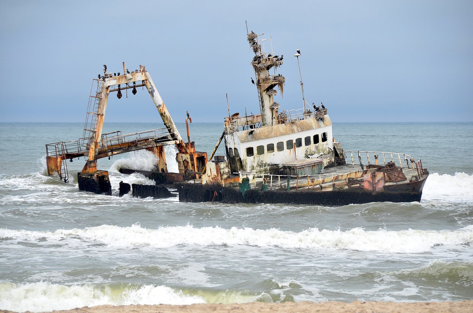 The rusted wreck of the trawler Zeila lying in grey surf off a sandy shore, its white wheelhouse, masts and winch frame still standing above the waves
