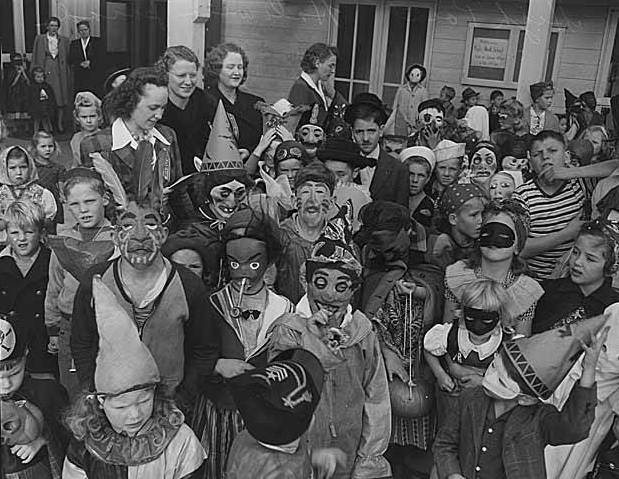 Black-and-white photograph of a crowd of children in homemade and mask-style Halloween costumes, with witch hats, paper masks and eye masks, and a few women standing behind them