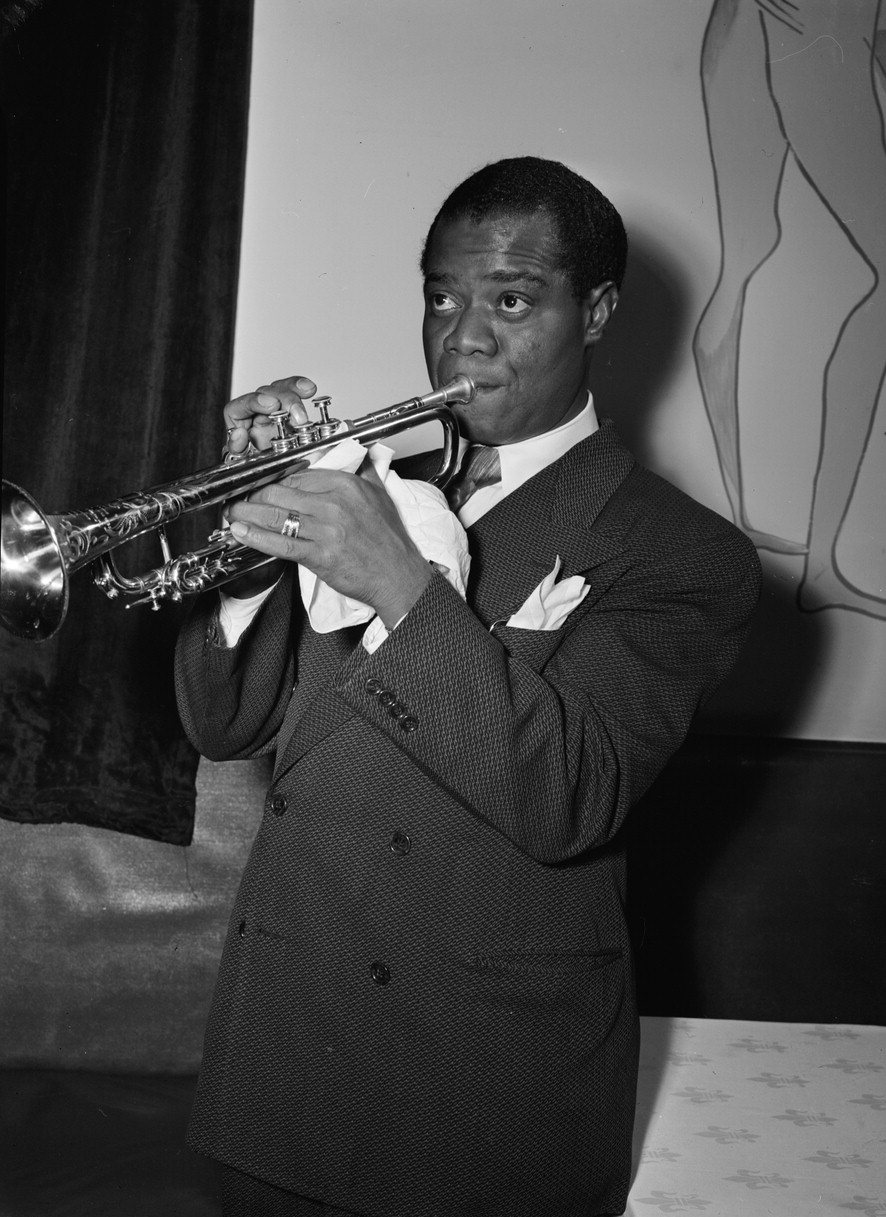 A black-and-white photograph of Louis Armstrong in a suit, playing the trumpet with a white handkerchief in his hand