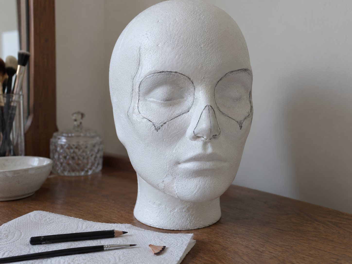 Illustration: Styrofoam mannequin head with a smooth white painted base and light black pencil outlines of wide eye sockets and a nose cavity, with a brow pencil and detail brush beside it