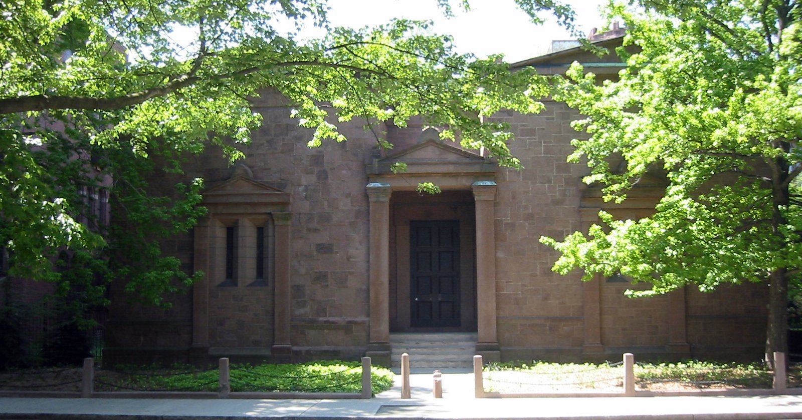 Front of the Skull and Bones Tomb on High Street in New Haven: a windowless brownstone building with a columned doorway and two narrow window frames, half hidden by leafy trees behind short stone posts