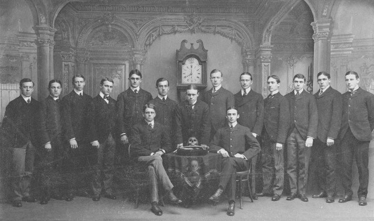 Group photograph of fifteen young men in dark suits, two seated beside a draped table holding a skull and crossed bones, the others standing in a row before a tall clock and a carved backdrop