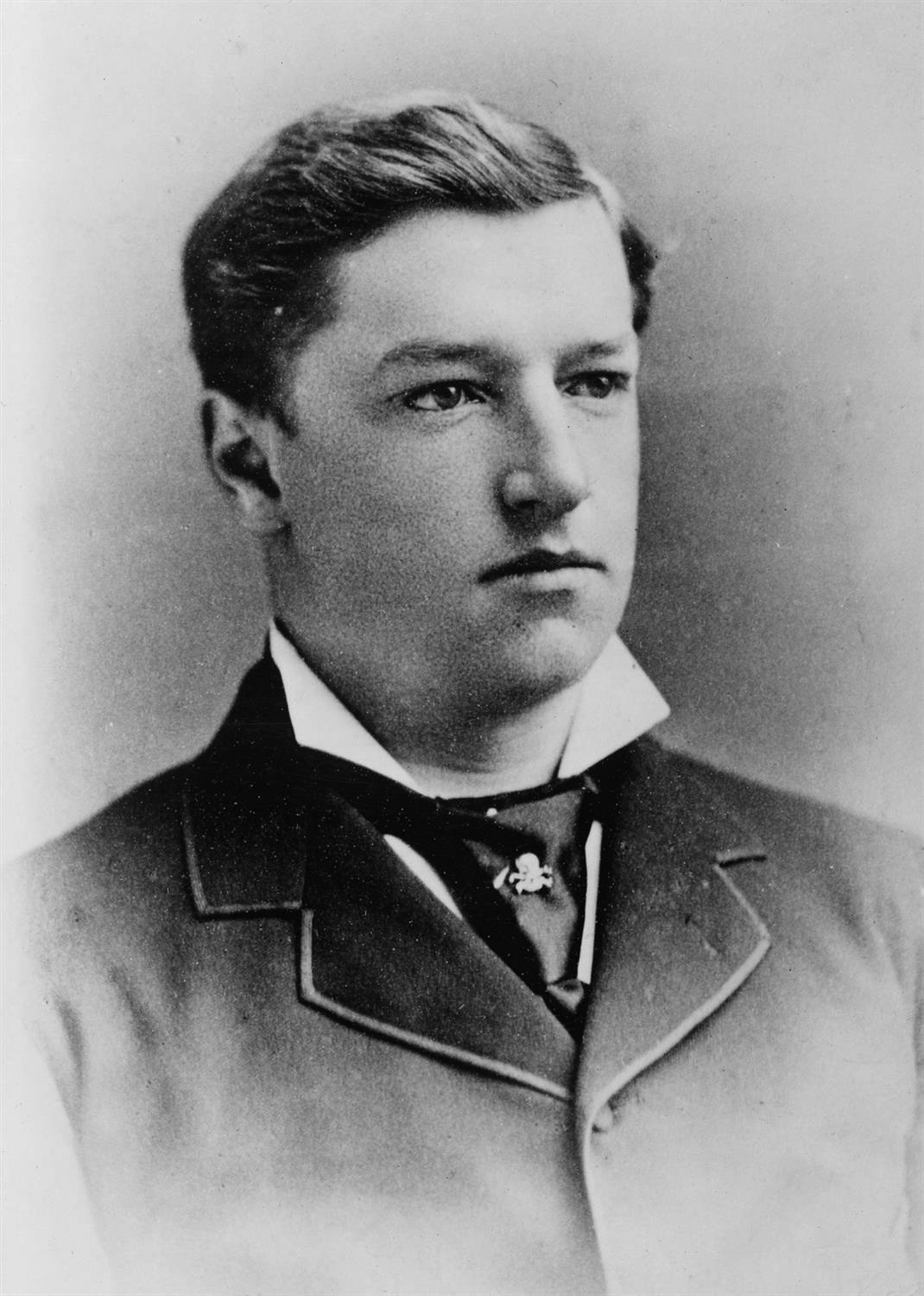 Black and white portrait of a young William Howard Taft with side-parted hair, in a jacket with a wide collar and a dark tie fastened with a small skull and crossbones pin