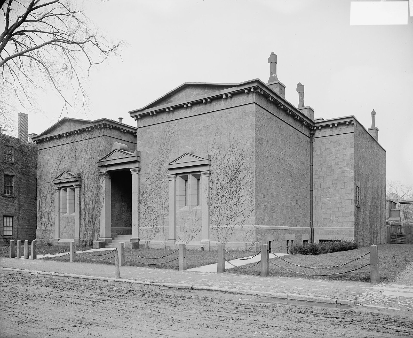 Black and white photograph of the Tomb from the street corner: a windowless stone hall with two pediments, a columned entrance, ivy on the walls and a low chain fence along a muddy road