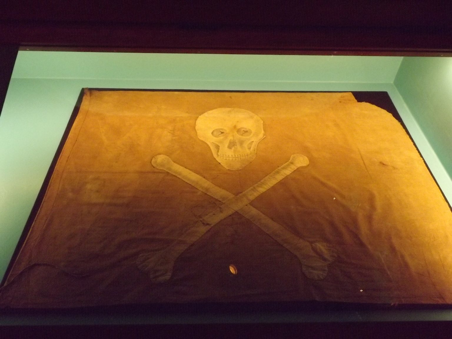 A faded orange-brown flag in a museum case, with a pale cloth skull and two crossed bones stitched to it
