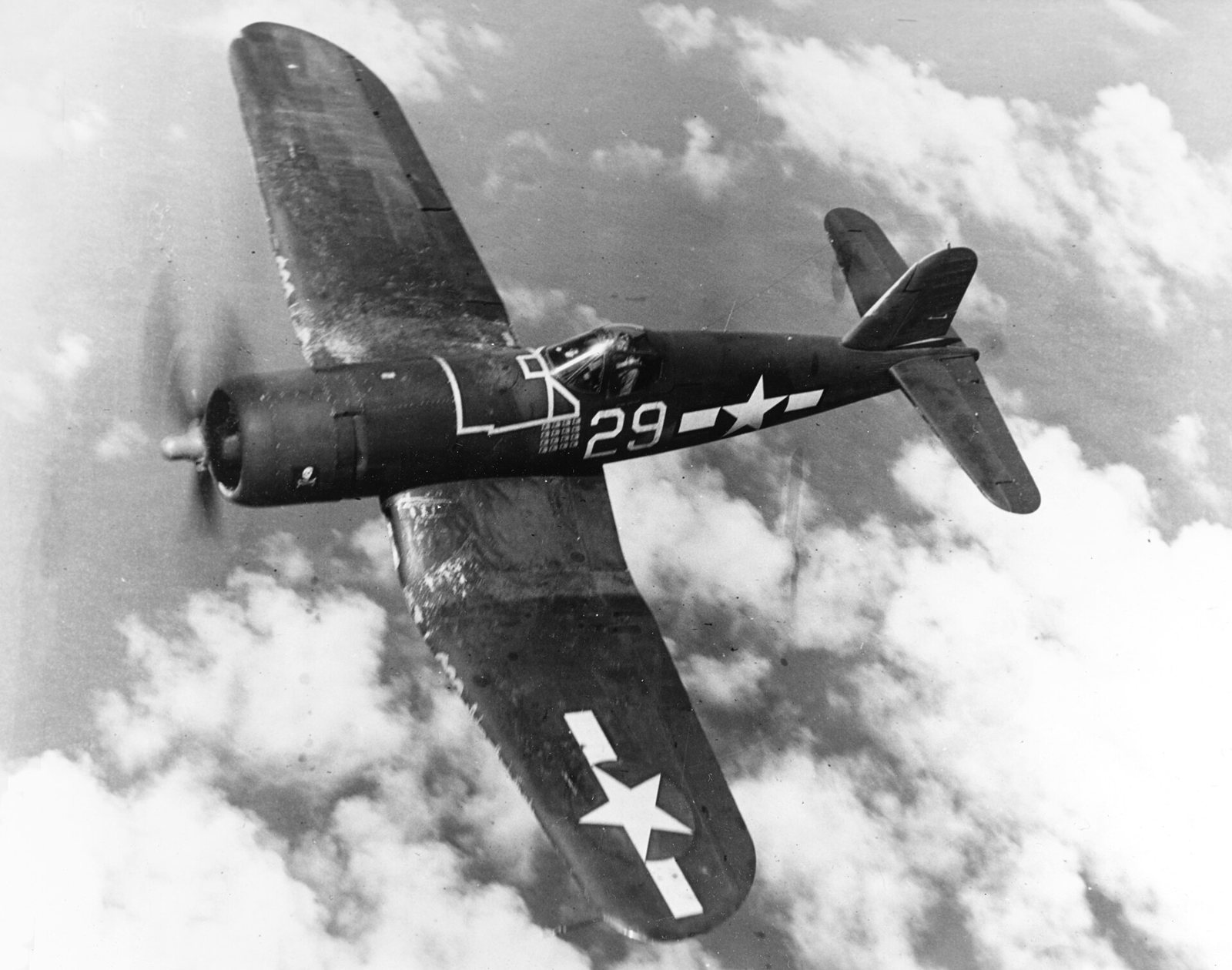 A dark gull-winged Navy fighter numbered 29 banking over clouds, with a small skull and crossbones painted near its propeller