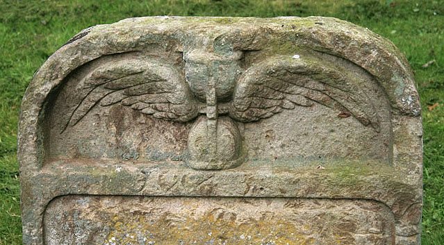 The top of a weathered headstone at Channelkirk Churchyard, carved with an hourglass spreading a pair of wings above its date