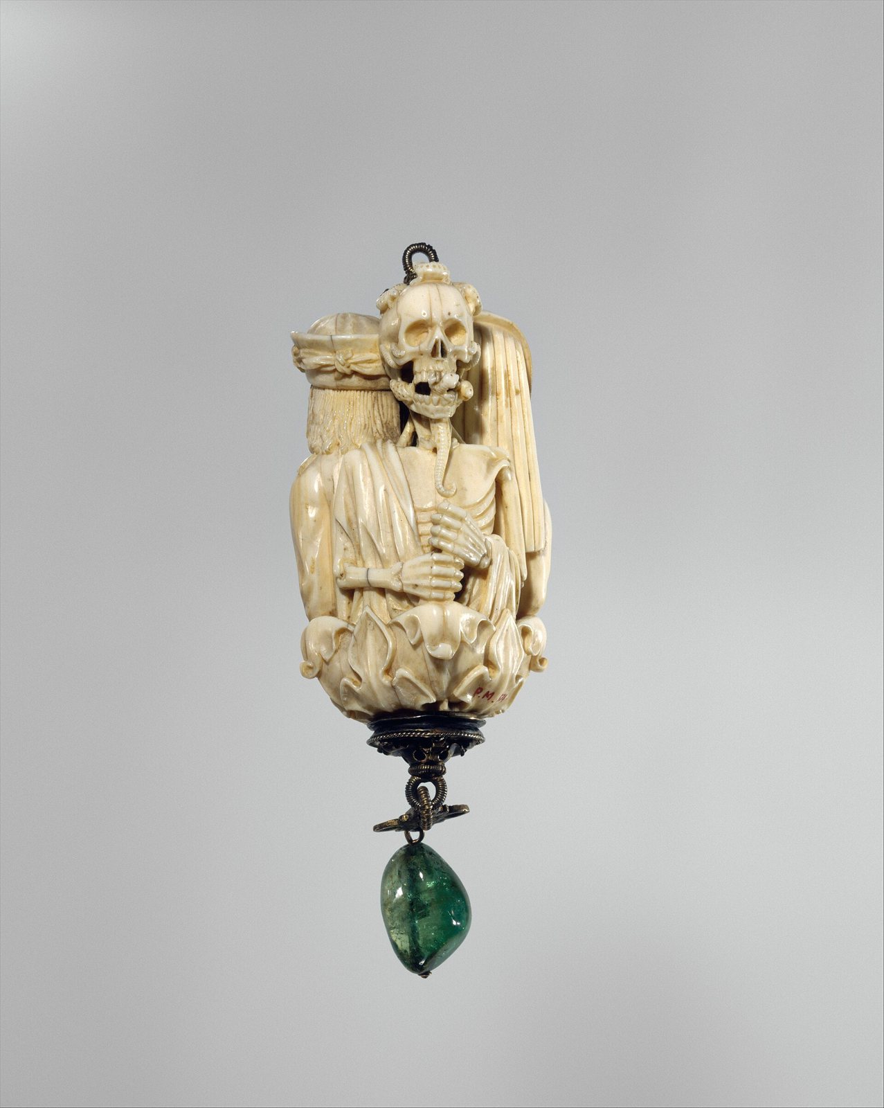 Carved ivory bead showing a skeletal figure with a grinning skull and a worm curling from its jaw, hanging above a green stone drop