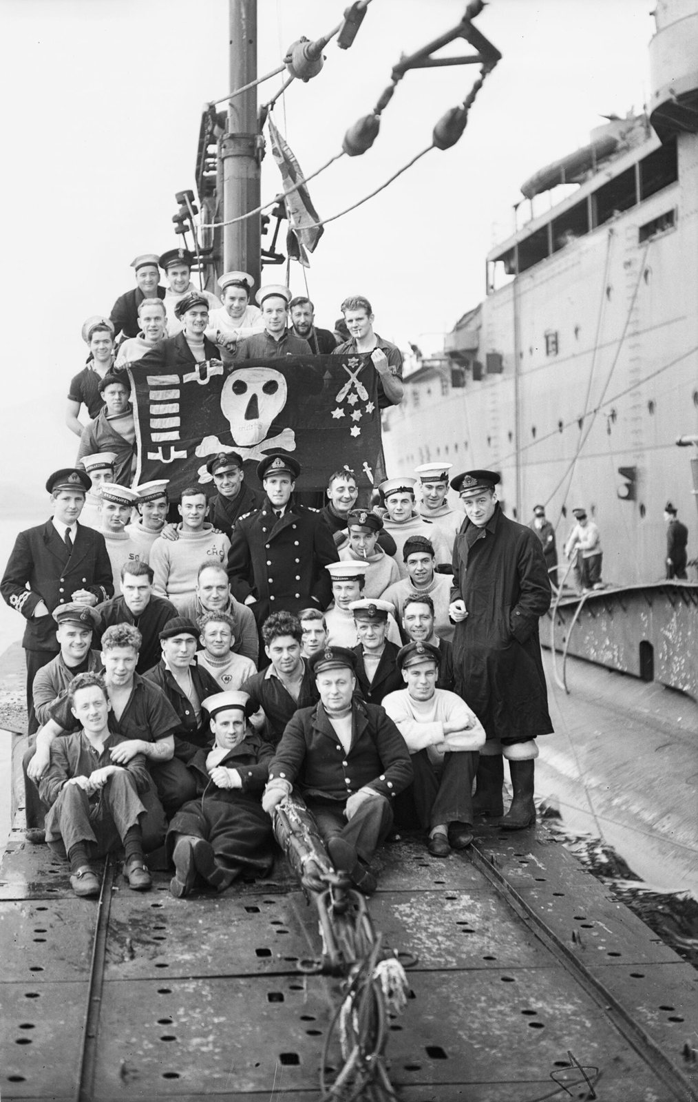 Fotografía en blanco y negro de la tripulación de un submarino reunida en cubierta junto a una gran bandera oscura con una calavera blanca, en 1942