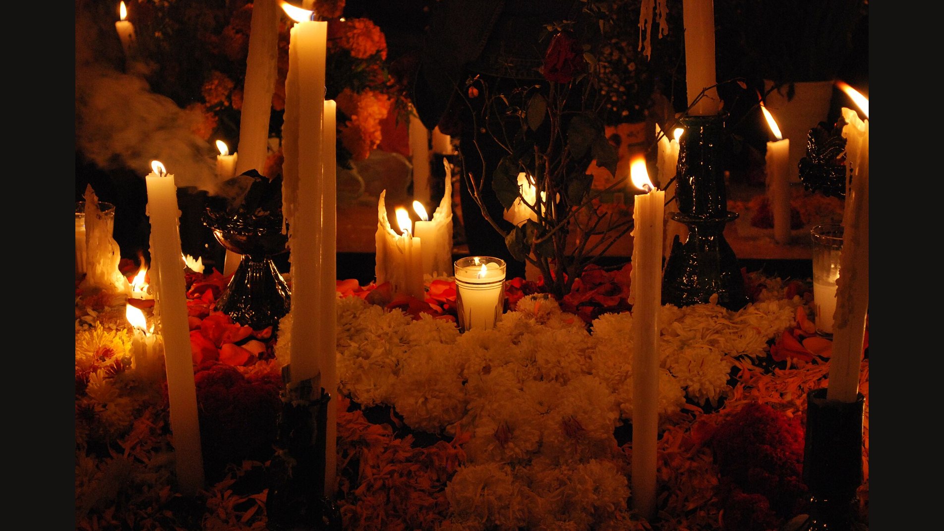 Tall white candles burning among heaps of white and orange marigolds on an ofrenda at night, with more flames glowing in the dark behind