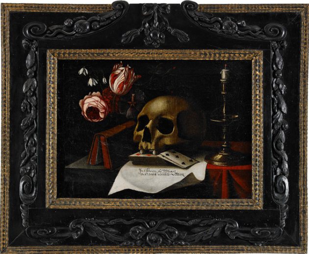 A dark still life in a carved black frame: a skull rests on a book and playing cards beside a brass candlestick, with a rose and a tulip behind it and a sheet of paper in front