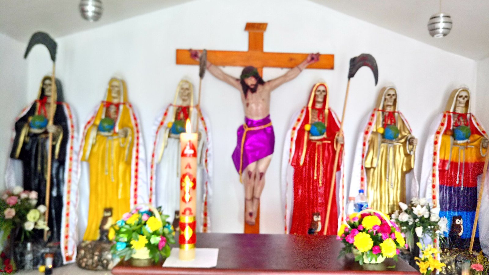 The main altar of a small temple: seven robed skeletal figures in black, yellow, white, red and striped robes stand around a crucifix, with flower arrangements and a tall decorated candle in front