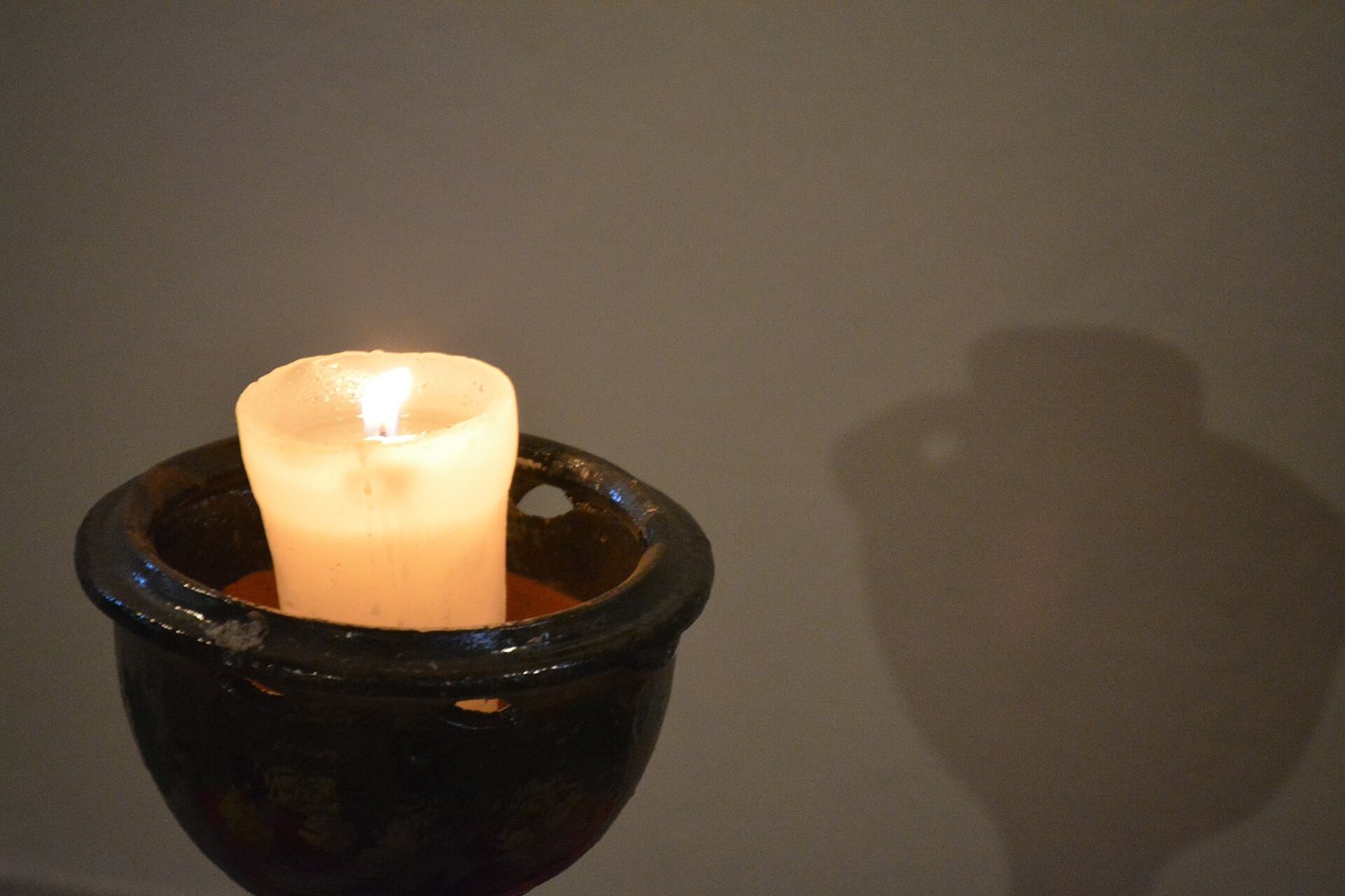 A single lit candle burns inside a dark glazed clay dish against a plain wall, casting a soft shadow behind it