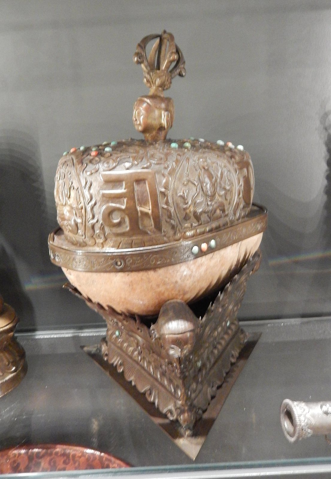 A Tibetan kapala in a display case: a bone bowl with an embossed metal lid set with turquoise and coral and topped by a vajra handle, resting on a triangular metal stand