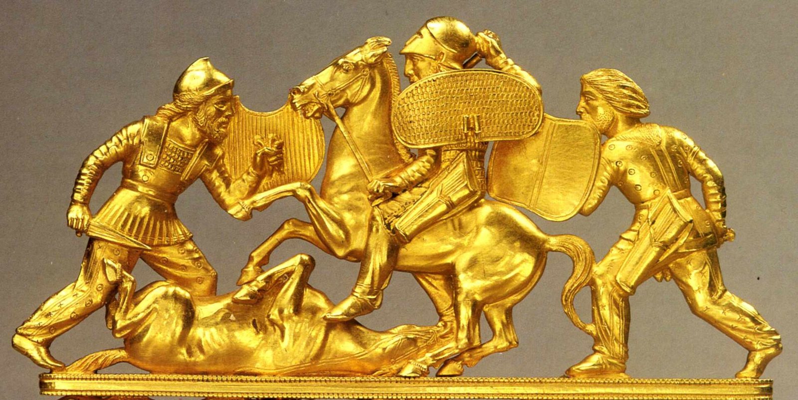 The gold top of a Scythian comb showing a battle: a helmeted rider on a rearing horse and two warriors on foot with shields, over a fallen horse