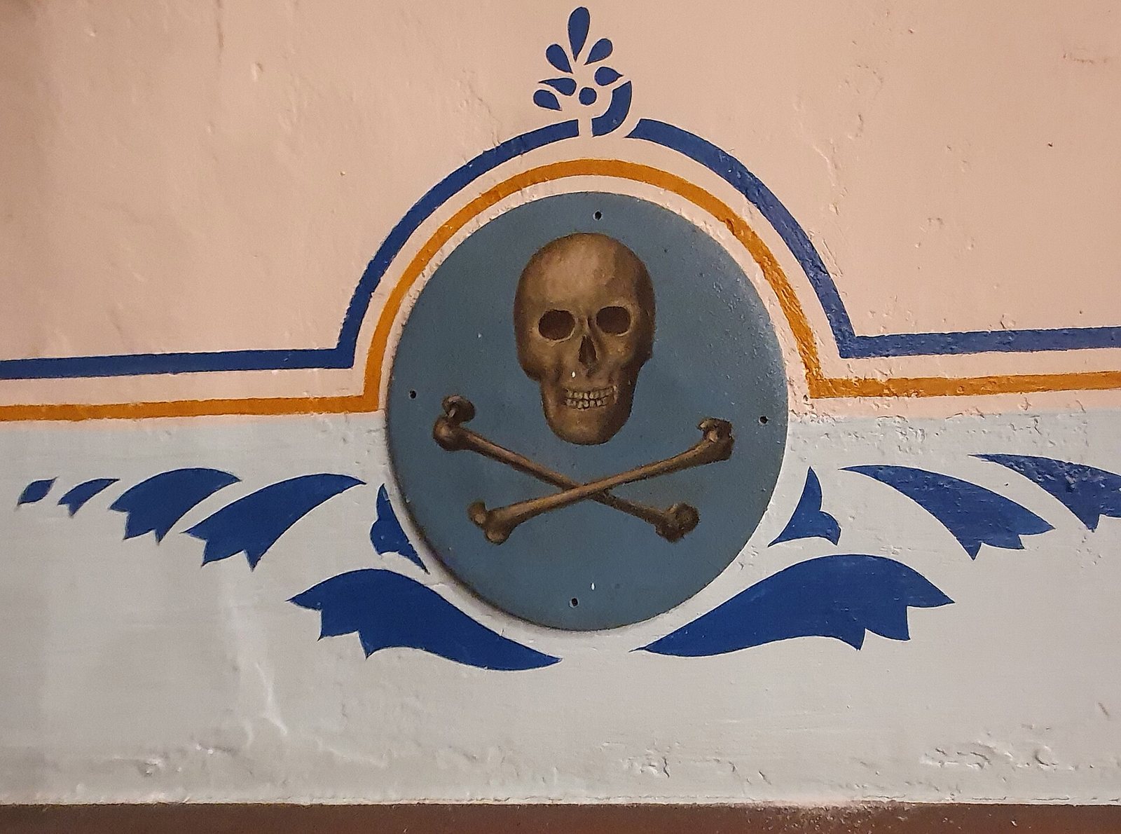 Painted roundel of a skull above crossed bones on a blue disc, set into a pink and pale blue wall with blue and orange decorative bands