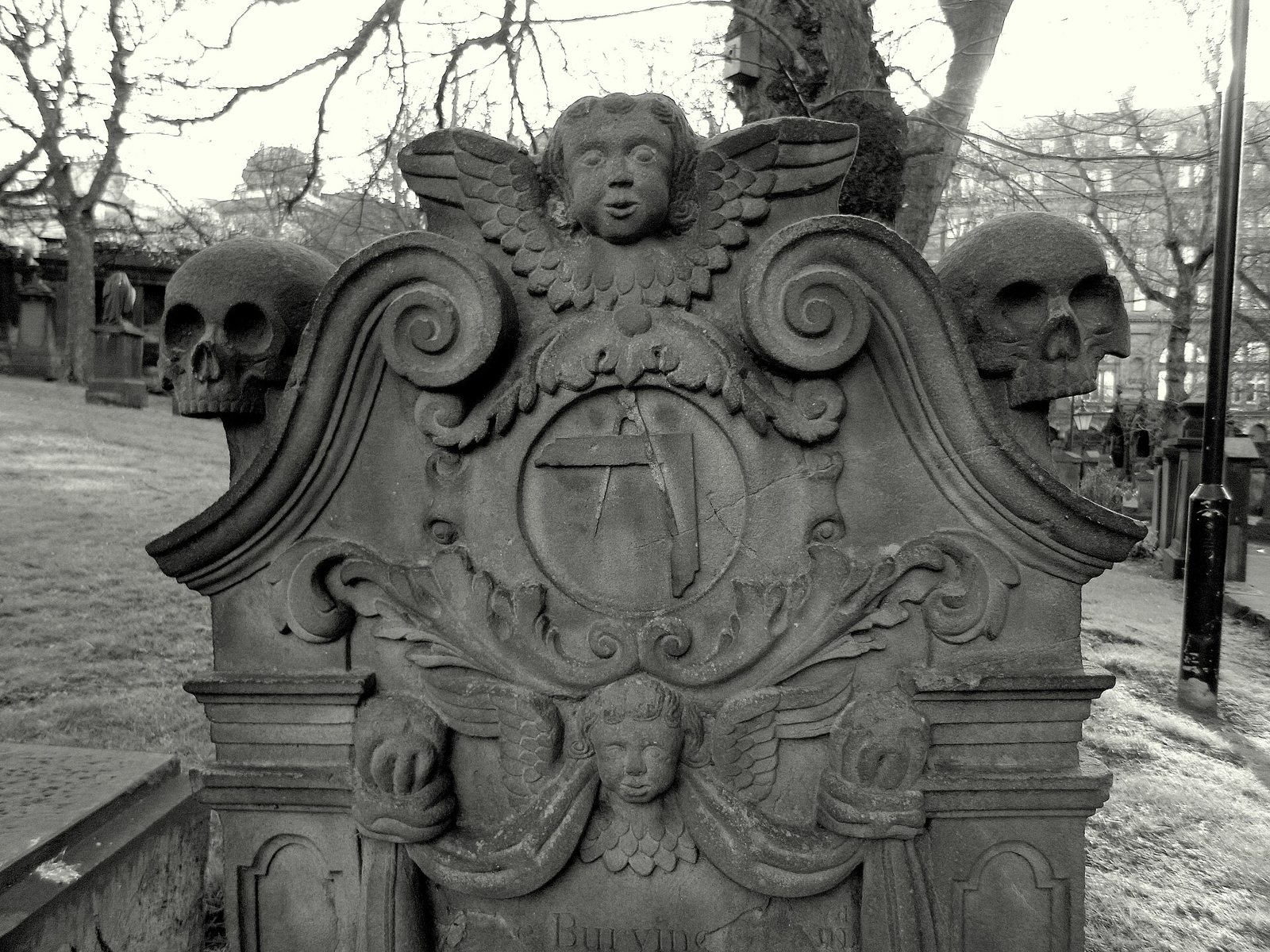 Black and white detail of a carved sandstone headstone in a churchyard: a square and compasses in a central roundel, framed by scrolls and two winged cherub heads, with a carved skull at each upper corner