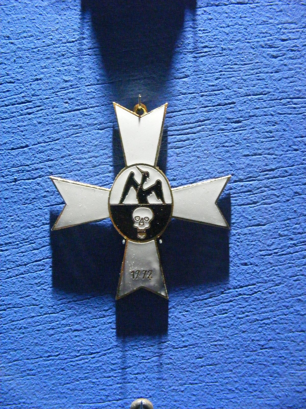 White enamel cross with flared arms hanging against a blue wall, its centre showing a black phoenix above a white skull, with the date 1772 on the lower arm