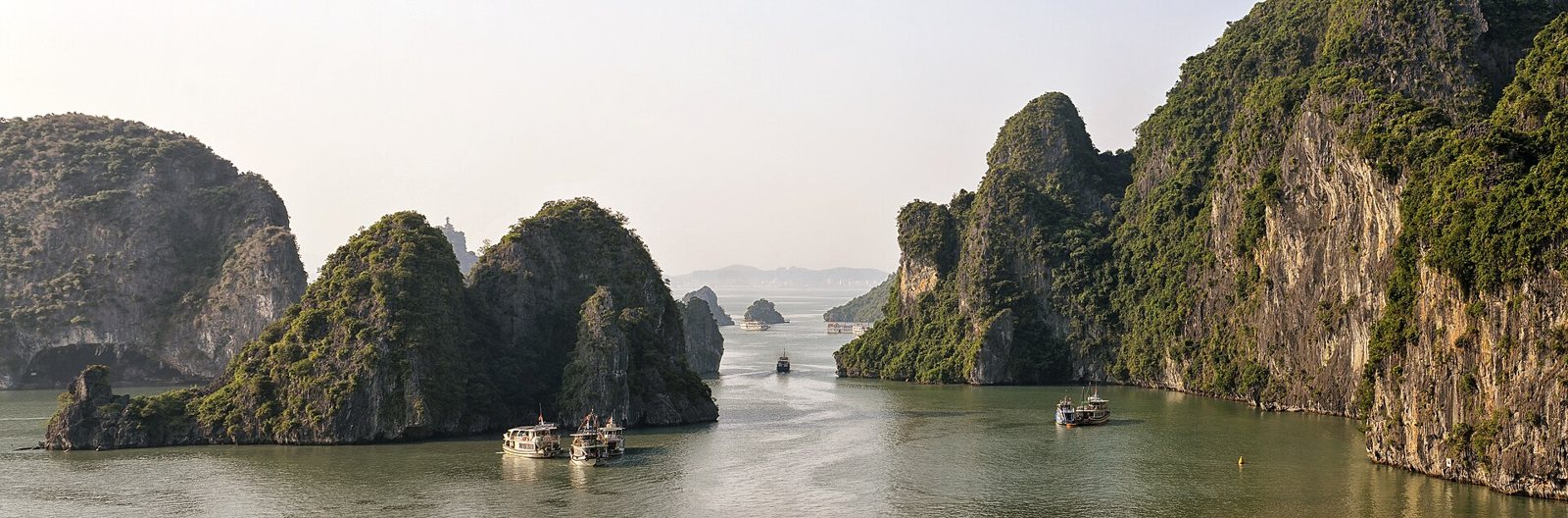 Limestone islands covered in green forest rising from the calm, grey-green water of Ha Long Bay in Vietnam, with small boats between them under a pale sky