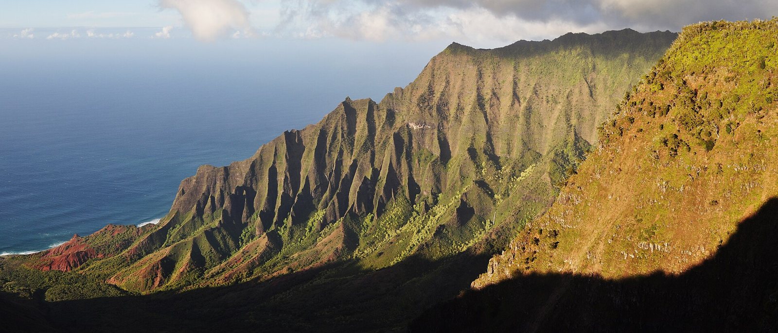 The steep, fluted green ridges of the Nā Pali coast on Kauaʻi in late sun, dropping to the blue Pacific on the left, with the Kalalau Valley in shadow below
