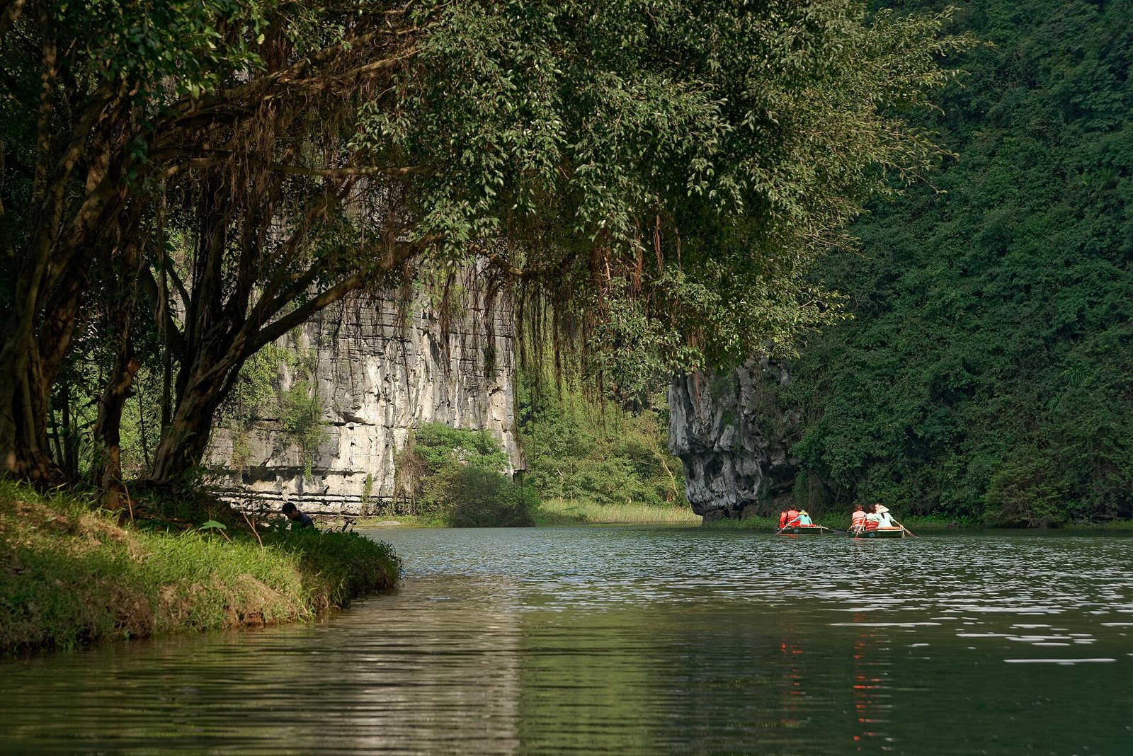 A still green river in the Tràng An area of Ninh Bình, Vietnam, with pale limestone cliffs and dense forest on the far bank and two small boats in the distance beneath overhanging trees