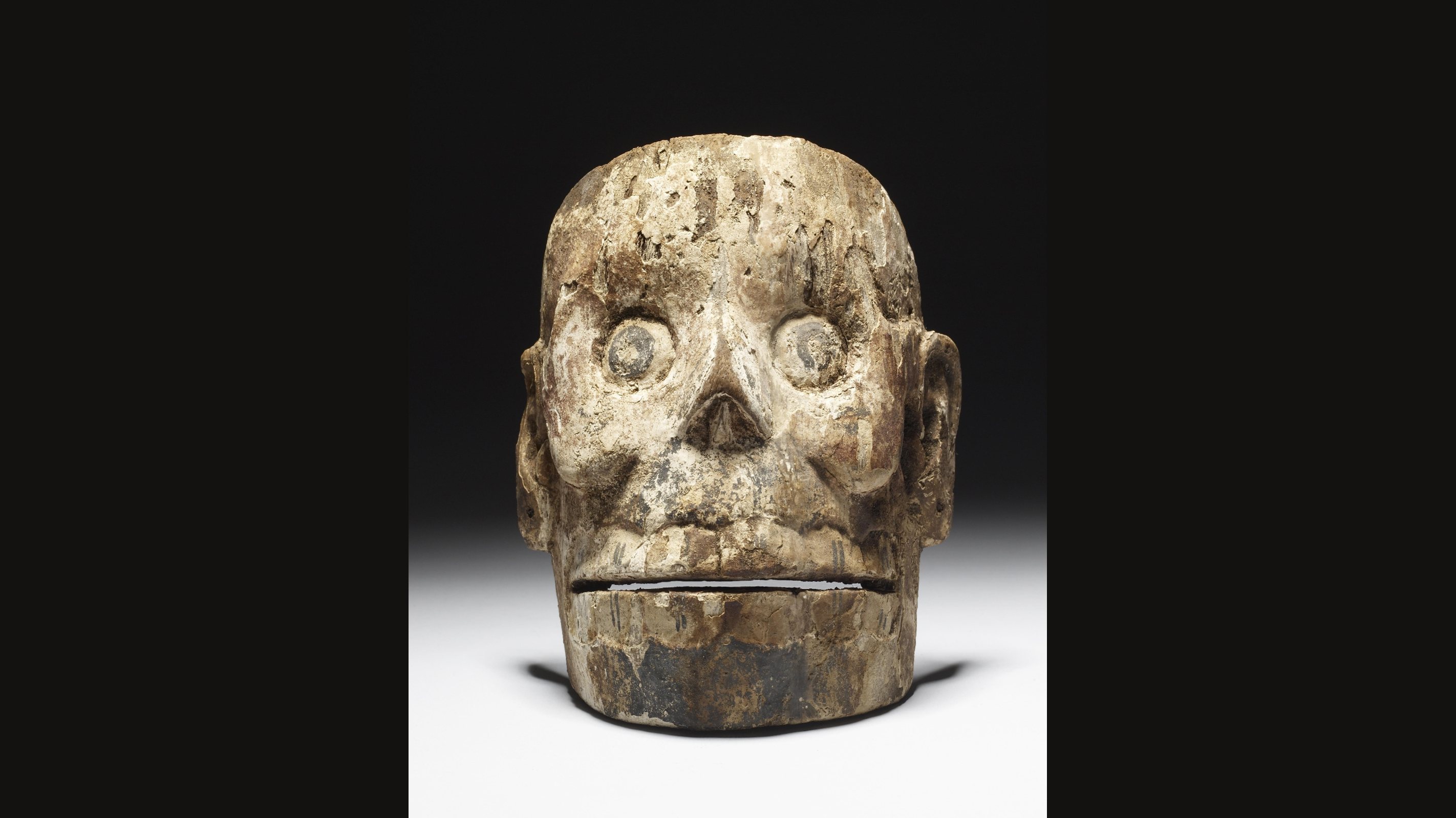 A weathered wooden skull mask with round hollows for eyes, a flat nose cavity and a closed jaw line, lit against a black background