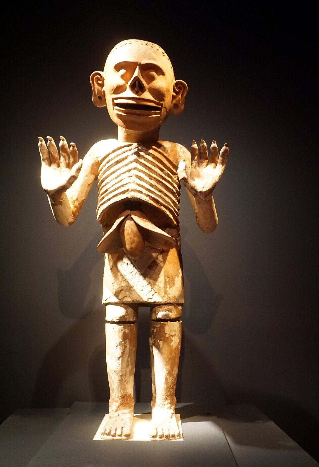 A tall standing clay figure of Mictlantecuhtli, painted white, with bare ribs, a skeletal grinning head and both hands raised, lit in a dark museum gallery