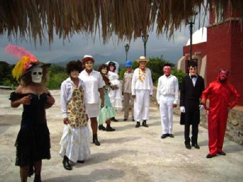 A group of Xantolo dancers in masks and costumes on a plaza in front of a red building: one in a black dress and plumed hat with a white skull mask, one in a red devil suit, and others in white clothes and masks