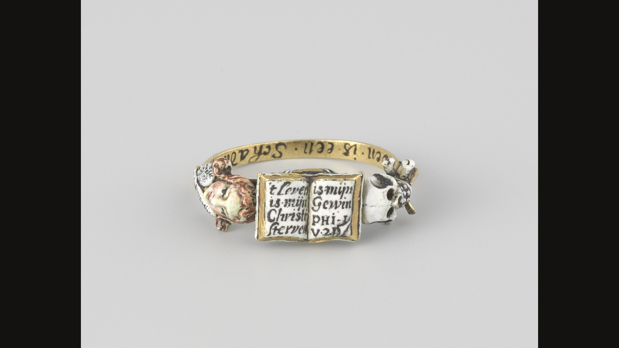 A gold memento mori ring with an open book of text on the bezel, a white enamel skull on one side and a small ornament on the other