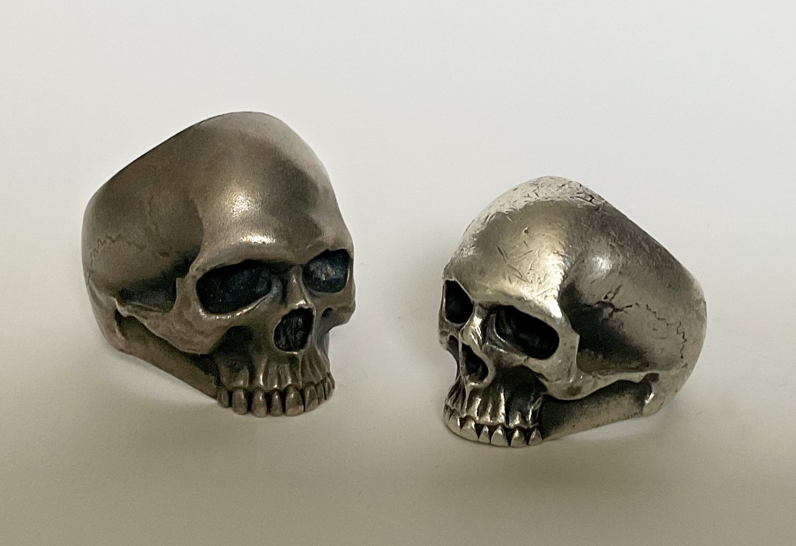 Two heavy silver skull rings side by side, one new and one worn smooth, on a pale ground