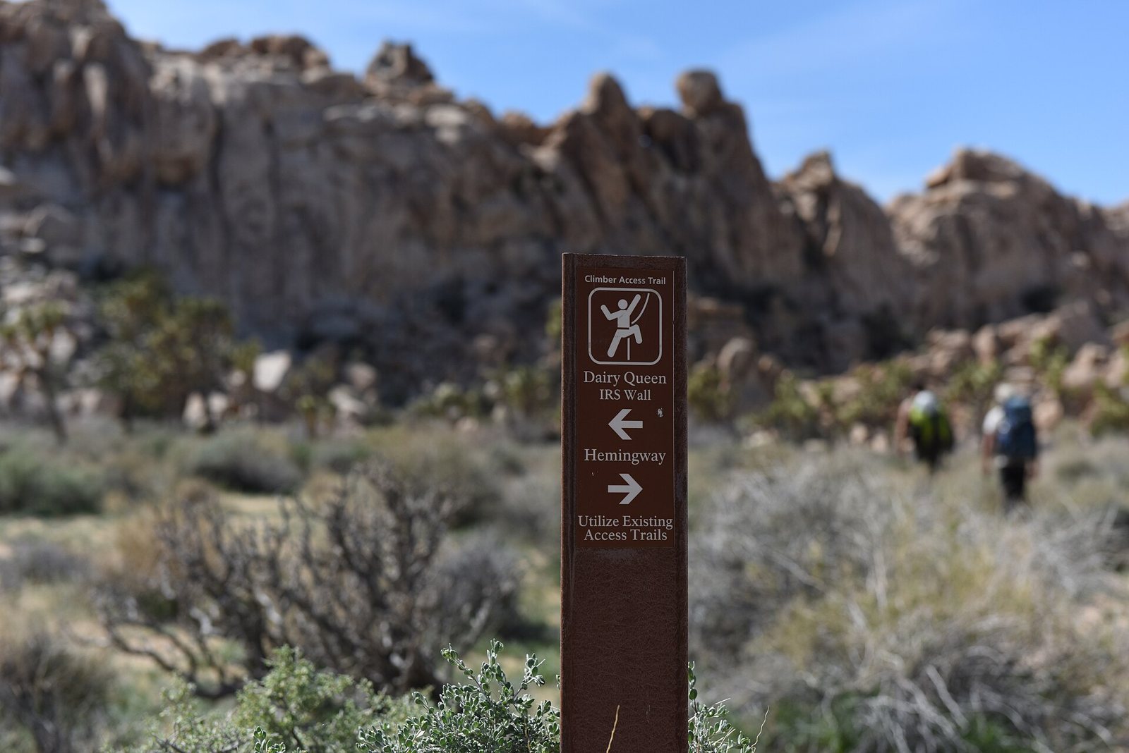 A brown National Park Service post with a climber pictogram and arrows pointing to named climbing areas, standing in desert scrub in front of a rocky outcrop, with two hikers with backpacks beyond it