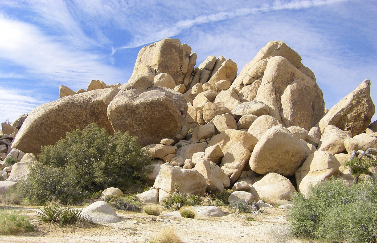 A heap of rounded monzogranite boulders in tan and cream at the entrance to Hidden Valley, with green desert shrubs at their base and wispy clouds above