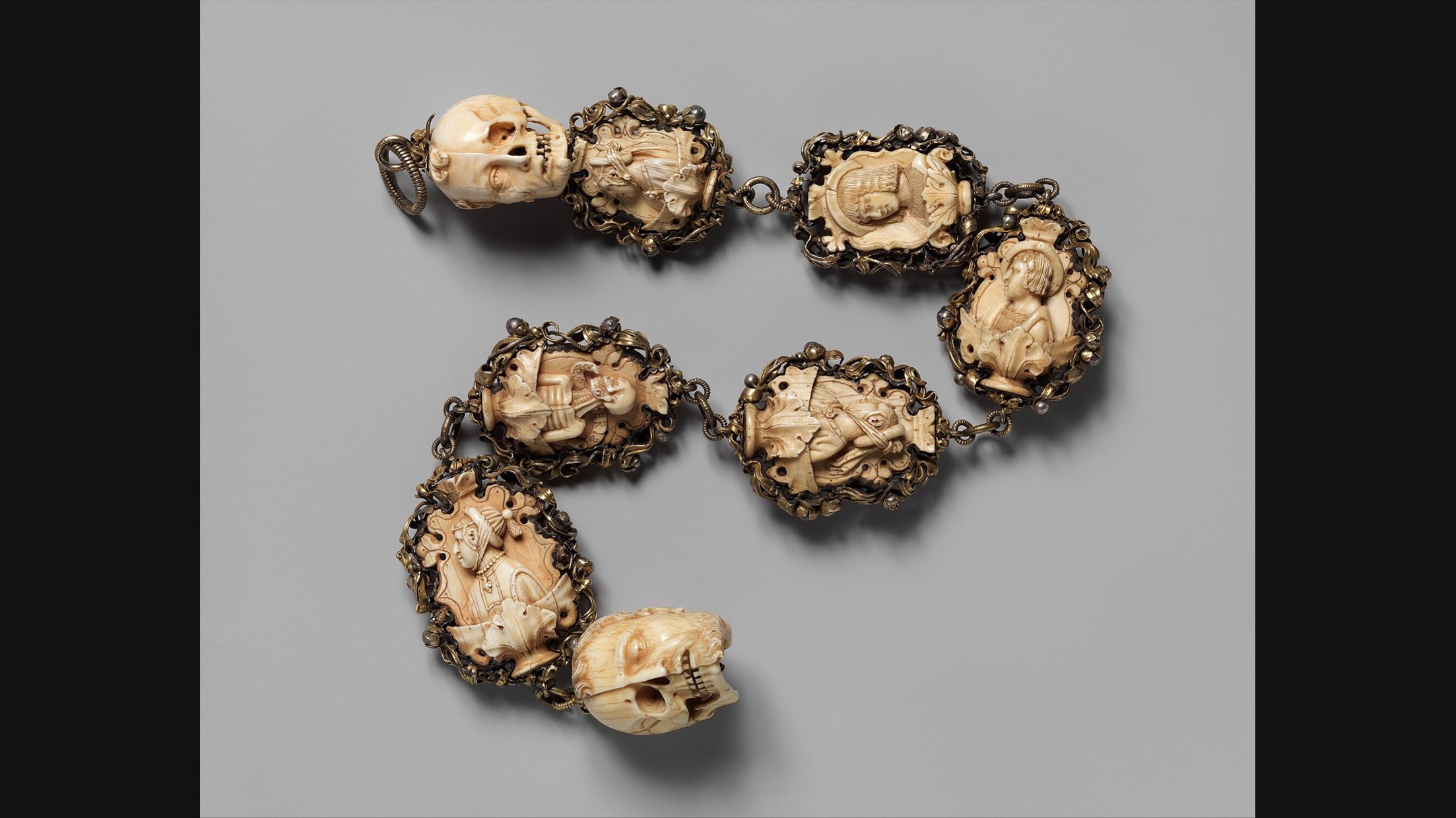 A German ivory rosary of about 1500 to 1525 laid out on grey: a carved skull at one end, links of carved ivory beads in gilt metal frames, and a second skull at the other end