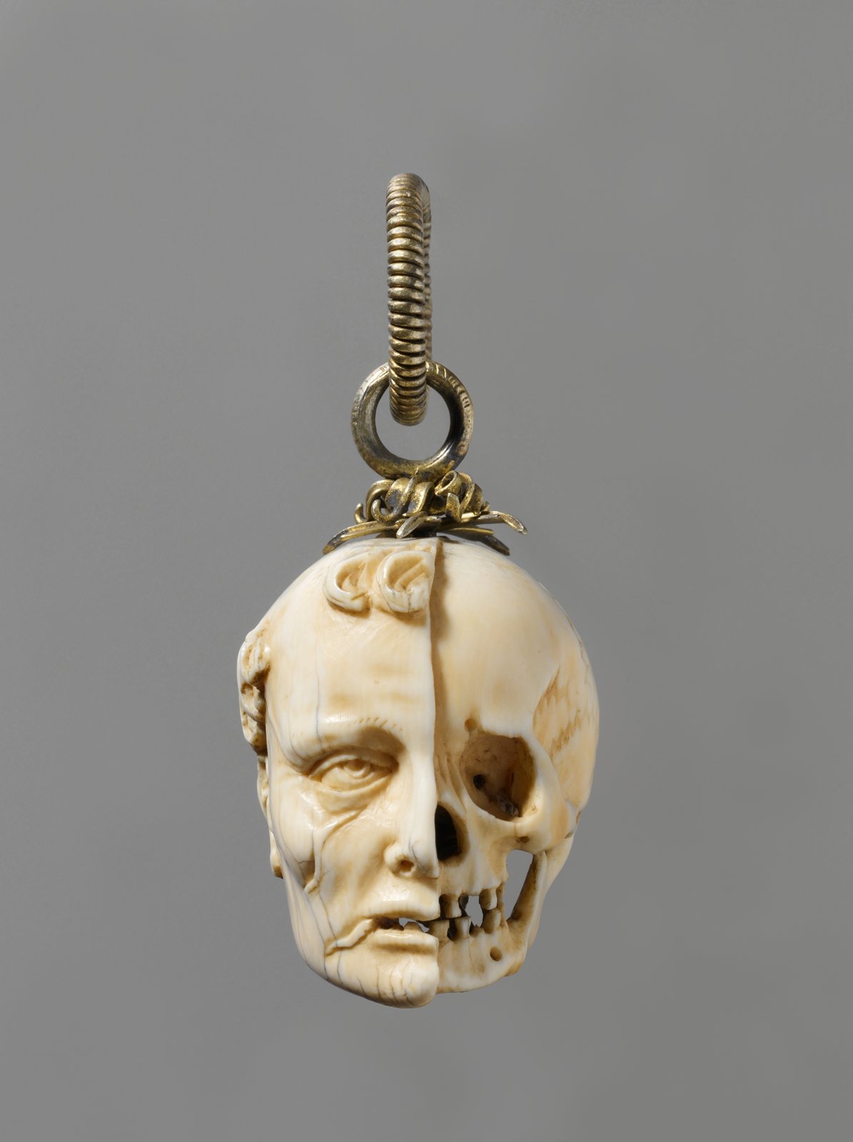 A carved ivory skull hanging from a small gilt ring, with half the face still fleshed and the other half bare bone