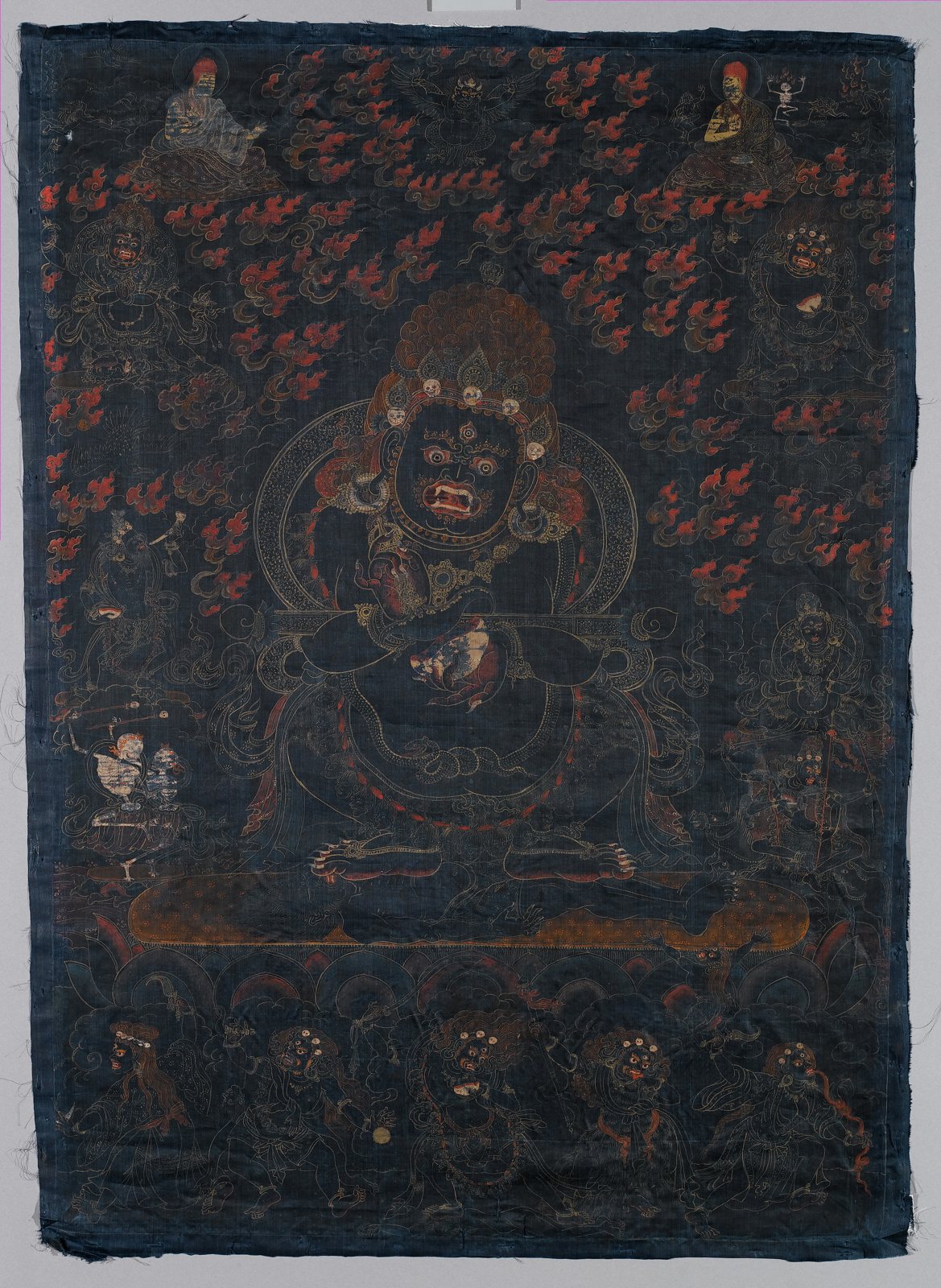 A dark Tibetan painting on blue silk of the wrathful protector Mahakala, a stout black figure with a crown of small white skulls, seated amid red flames and surrounded by smaller deities