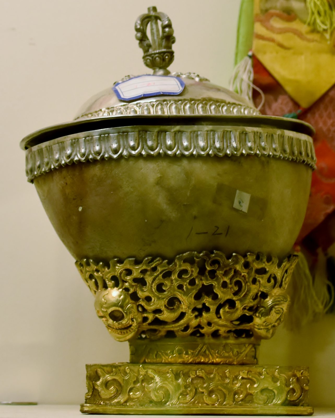 A skull cup with a domed silver lid topped by a vajra handle, set in an ornate pierced gilt stand with small skull faces on its rim, in a display case
