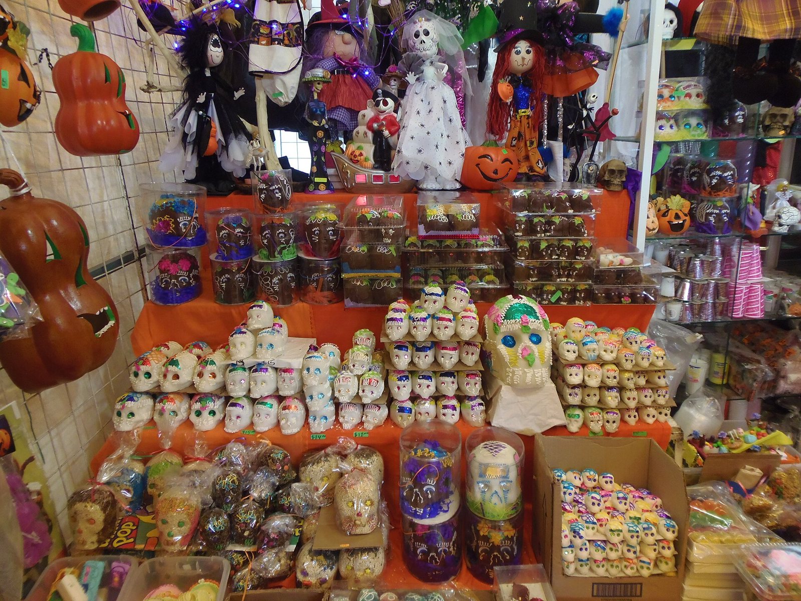 A market stall piled with small white sugar skulls decorated in bright icing and foil, with chocolate skulls above and Halloween decorations behind