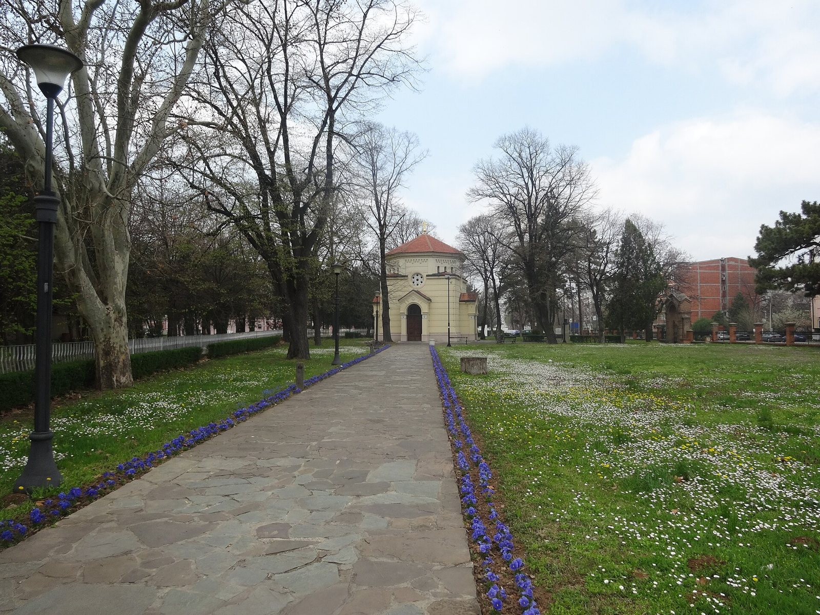 A paved path lined with flowers and bare trees leading through a park to the small chapel that encloses the Skull Tower