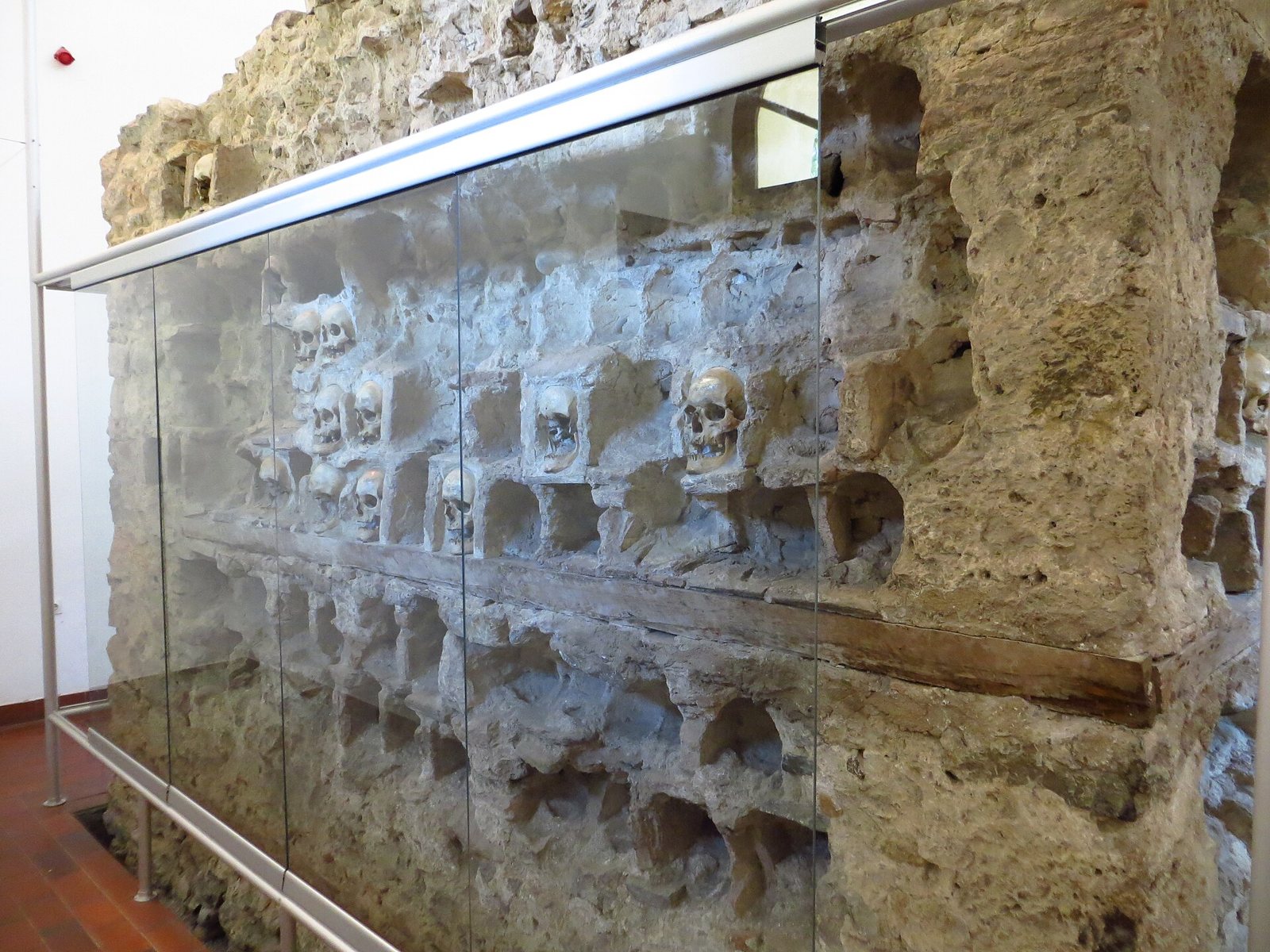 The surviving wall of the Skull Tower inside its chapel, behind a glass screen, with rows of square niches, a few still holding skulls